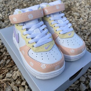 May include: White and pastel-colored sneakers with daisy accents. The shoes feature a white base with light yellow, peach, and lavender details. The straps and toe area are adorned with small daisy patterns. The sneakers are sitting on a gray box.