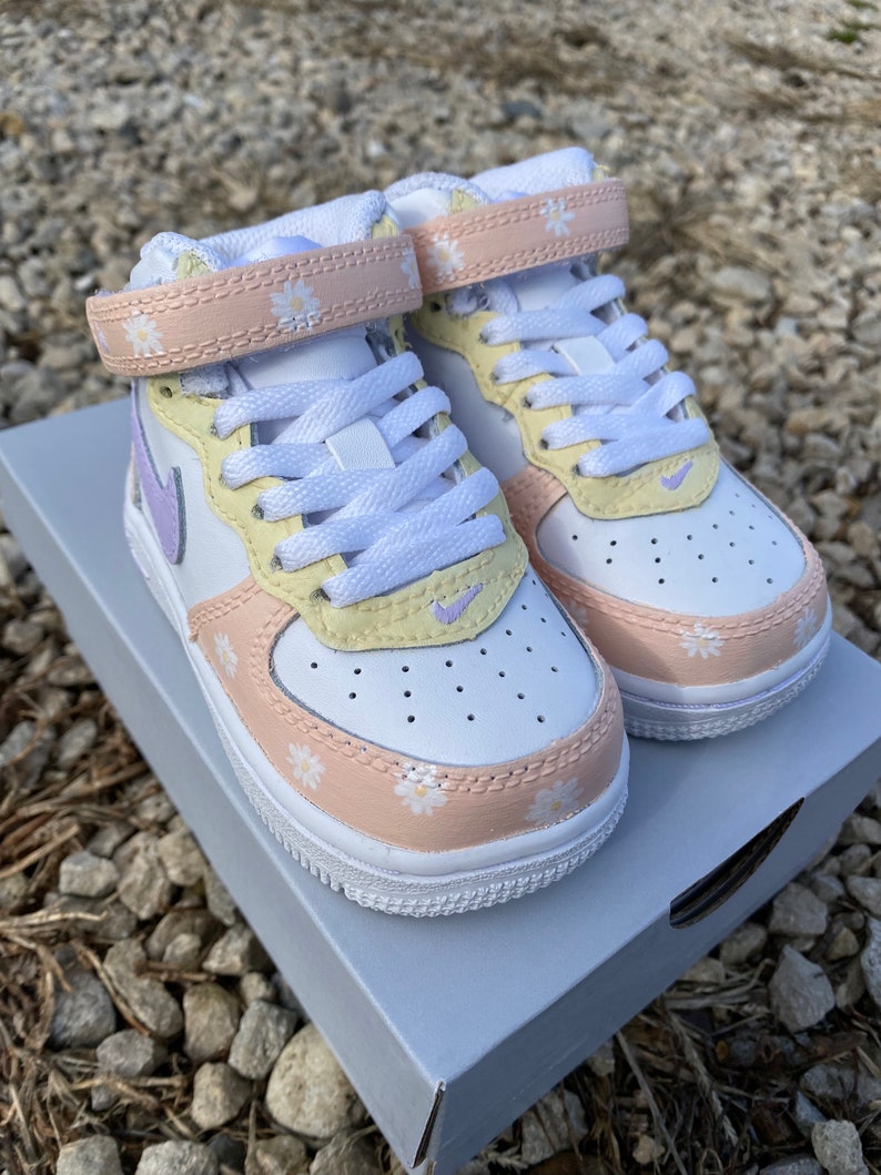 May include: White and pastel-colored sneakers with daisy accents. The shoes feature a white base with light yellow, peach, and lavender details. The straps and toe area are adorned with small daisy patterns. The sneakers are sitting on a gray box.