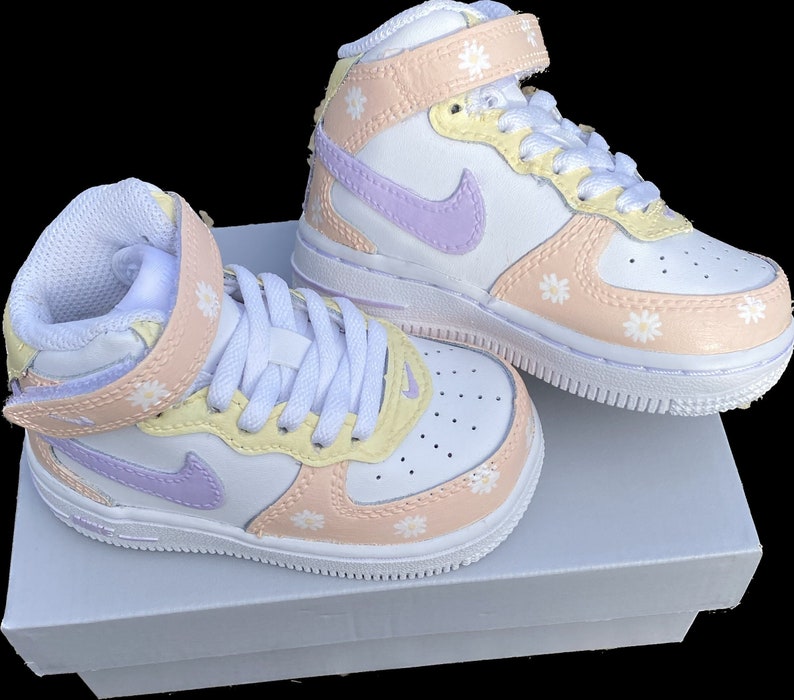 May include: A pair of white, high-top sneakers with pastel accents. The shoes feature a light purple Nike swoosh, peach and yellow panels, and small white daisy designs. The sneakers have white laces and a velcro strap. The shoes are on a white box.