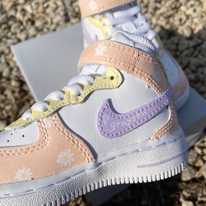 May include: White and pastel-colored high-top sneakers with daisy accents. The sneakers feature a white base with light pink, yellow, and lavender details. The Nike swoosh is lavender with the name "Grayson" written on it.