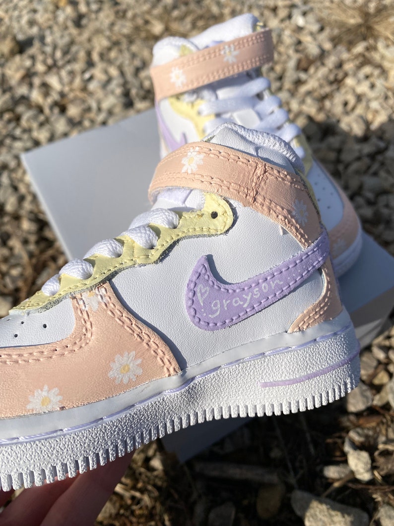 May include: White and pastel-colored high-top sneakers with daisy accents. The sneakers feature a white base with light pink, yellow, and lavender details. The Nike swoosh is lavender with the name "Grayson" written on it.