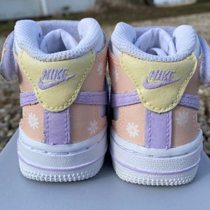 May include: A pair of baby sneakers with a pastel color scheme. The shoes feature a white sole, a light peach body, and accents of yellow and lavender. White daisy flower patterns are scattered across the sides. The Nike logo is visible on the heel.