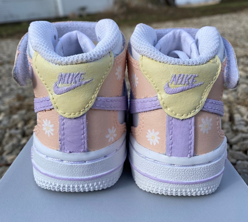 May include: A pair of baby sneakers with a pastel color scheme. The shoes feature a white sole, a light peach body, and accents of yellow and lavender. White daisy flower patterns are scattered across the sides. The Nike logo is visible on the heel.