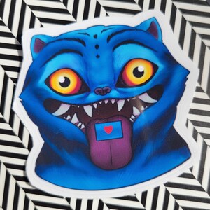 Op de afbeelding: Een blauwe kattensticker met grote gele ogen, scherpe tanden en een paarse tong. De tong heeft een blauwe rechthoek met een rood hart. De sticker staat op een zwart-wit gestreepte achtergrond.