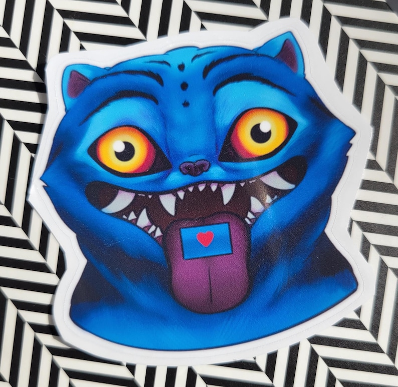 Op de afbeelding: Een blauwe kattensticker met grote gele ogen, scherpe tanden en een paarse tong. De tong heeft een blauwe rechthoek met een rood hart. De sticker staat op een zwart-wit gestreepte achtergrond.