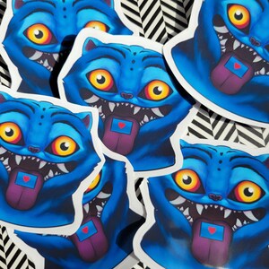 Op de afbeelding: Een verzameling blauwe kattenstickers met open monden, scherpe tanden en uitgestoken tongen. De tongen zijn paars met een kleine blauwe rechthoek en een rood hart. De katten hebben gele en oranje ogen.