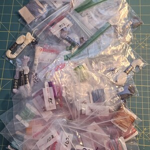 May include: A collection of embroidery floss skeins in various colours, each individually packaged in clear plastic bags. Many bags feature white paper labels with handwritten colour numbers, including 95, 103, 3072, 3823, and 451, organised for craft projects.