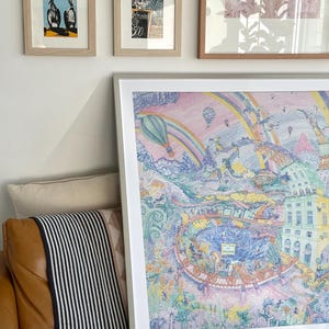 Könnte beinhalten: Ein großes, gerahmtes, farbenfrohes Kunstwerk mit einem skurrilen Stadtlandschaftsdesign, das Heißluftballons, einen Regenbogen und Gebäude zeigt. Zwei kleinere gerahmte Drucke von Vögeln und einem abstrakten Design befinden sich darüber. Das Kunstwerk lehnt an einem braunen Ledersofa.