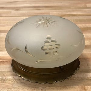 May include: Frosted glass ceiling light fixture with a starburst design and etched grape cluster. The light sits on a brass-colored scalloped base. The glass dome is a soft white color.