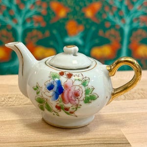 May include: A small, white porcelain teapot with a gold handle and spout. The teapot is decorated with a floral design featuring blue, pink, and red flowers with green leaves. The lid is topped with a gold accent.