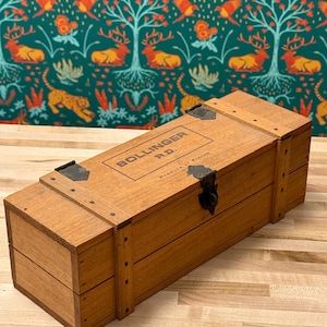 May include: A rectangular wooden box with metal hinges and a clasp. The box is light brown with the text "BOLLINGER R.D." printed on the top. The box is sitting on a light brown wooden surface.