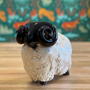 May include: A ceramic ram figurine with a black glossy head and horns, and a textured, off-white body. The ram has small black feet and is set against a patterned background. The figurine is a decorative item.