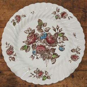 Puede incluir: Plato de cerámica blanca con borde ondulado y diseño floral. El plato presenta un ramo central de rosas rojas y marrones con detalles azules y verdes. Arreglos florales más pequeños adornan el borde.