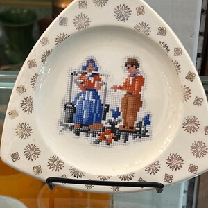 May include: A white ceramic triangular plate with a gold and brown floral pattern around the edge. The center of the plate features a cross-stitch design of two figures in traditional clothing.