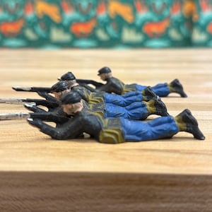 Puede incluir: Soldados de juguete en miniatura en posición de disparo tendida. Las figuras están pintadas con chaquetas negras, pantalones azules y sombreros negros. Los soldados están tumbados sobre una superficie de madera. El fondo es una tela estampada con colores verdes, naranjas y amarillos.