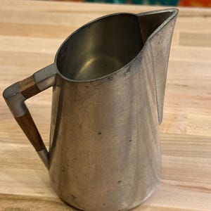 Puede incluir: Una jarra de metal vintage con un asa de madera. La jarra tiene un acabado plateado y un pico para verter fácilmente. El asa es de madera y está sujeta a la jarra con soportes metálicos. La jarra probablemente se utiliza para servir bebidas.