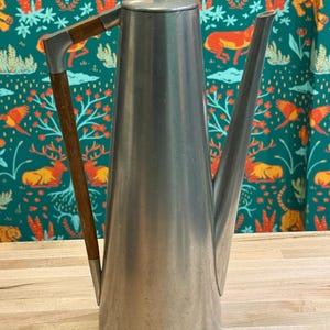 Puede incluir: Una cafetera alta de metal cepillado con asa y pomo de madera. La cafetera tiene un pico largo y delgado y un diseño minimalista. El asa es de madera oscura y está unida con soportes metálicos. El fondo presenta una tela estampada color turquesa.