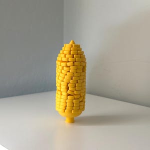 May include: A bright yellow, three-dimensional corn cob figurine featuring a textured surface composed of numerous small, rectangular, brick-like segments arranged in a vertical pattern, tapering to a point at the top and a small base.