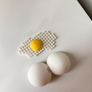 May include: A white plastic egg-shaped container with a visible seam and a separate flat, white, grid-patterned piece featuring a raised, circular, yellow centre, resembling a fried egg, displayed on a plain white surface.