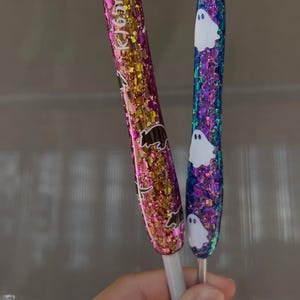 K&ouml;nnte beinhalten: Zwei Glitzerstifte. Einer ist rosa und goldfarben mit einem B&auml;renmuster, der andere ist blau und lila mit wei&szlig;en Geistermustern. Die Stifte haben wei&szlig;e Basen und werden in einer Hand gehalten.