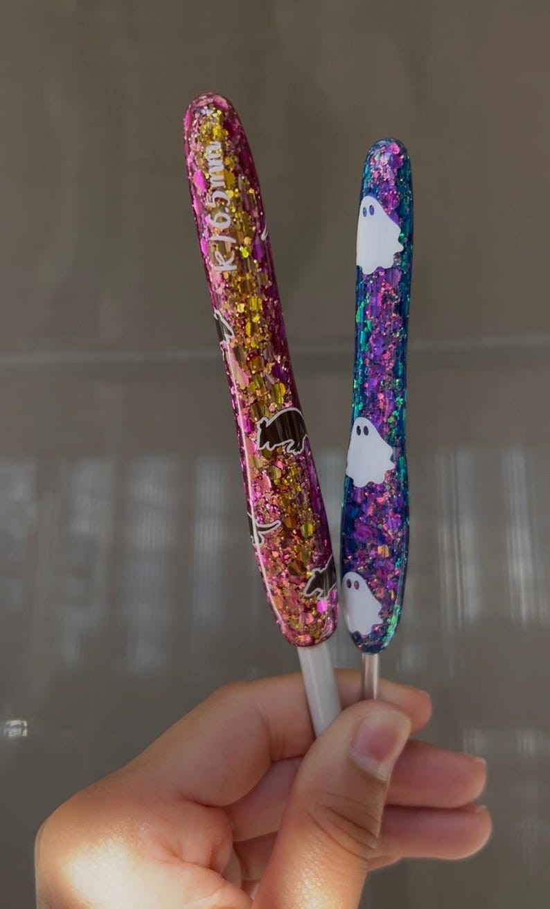 K&ouml;nnte beinhalten: Zwei Glitzerstifte. Einer ist rosa und goldfarben mit einem B&auml;renmuster, der andere ist blau und lila mit wei&szlig;en Geistermustern. Die Stifte haben wei&szlig;e Basen und werden in einer Hand gehalten.