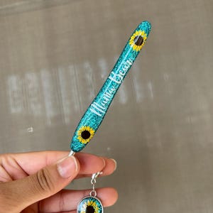 K&ouml;nnte beinhalten: Ein t&uuml;rkisfarbener Glitzerstift mit dem wei&szlig;en Schriftzug "Mama Bear". Der Stift hat Sonnenblumenmuster und einen passenden Anh&auml;nger mit einem Sonnenblumenbild. Der Anh&auml;nger ist mit einem silbernen Verschluss am Stift befestigt.