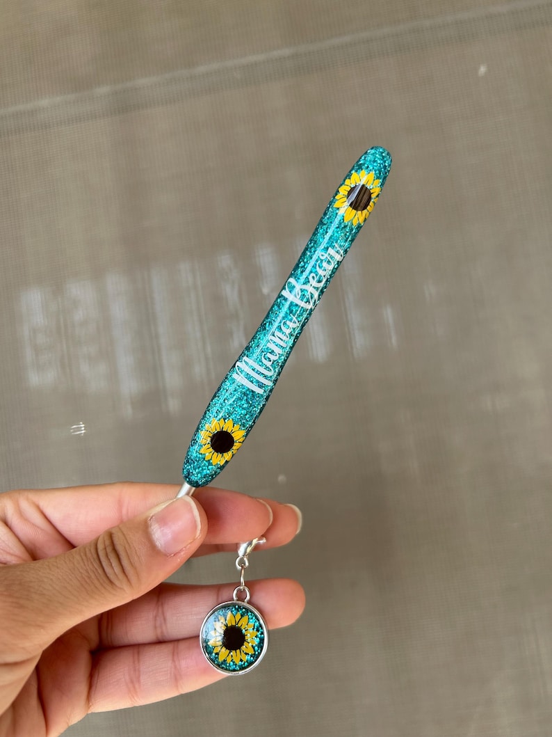 K&ouml;nnte beinhalten: Ein t&uuml;rkisfarbener Glitzerstift mit dem wei&szlig;en Schriftzug "Mama Bear". Der Stift hat Sonnenblumenmuster und einen passenden Anh&auml;nger mit einem Sonnenblumenbild. Der Anh&auml;nger ist mit einem silbernen Verschluss am Stift befestigt.