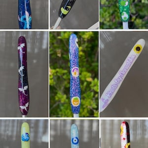 K&ouml;nnte beinhalten: Eine Sammlung dekorativer H&auml;kelnadeln mit verschiedenen Designs. Die H&auml;kelnadeln weisen Glitzer und Charakterdesigns auf, darunter Stitch, Yoshi, Halloween-Elemente, Drachen und Sonnenblumen. Jede H&auml;kelnadel ist ein einzigartiges, farbenfrohes Handwerkzeug.