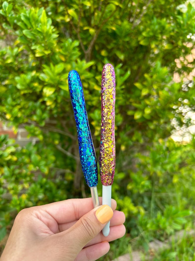 Peut inclure: Deux outils pour les ongles recouverts de paillettes, l'un bleu et l'autre dor&eacute;, sont pr&eacute;sent&eacute;s sur un fond vert feuillu. Les outils ont une poign&eacute;e argent&eacute;e et une base blanche. L'outil bleu a une finition paillet&eacute;e bleu sarcelle et bleu, tandis que l'outil dor&eacute; a une finition paillet&eacute;e or, rose et jaune.