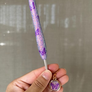 K&ouml;nnte beinhalten: Ein lila H&auml;kelhaken und ein passender Ohrring. Der Haken ist mit Glitzer und Schmetterlingsmotiven bedeckt. Der Ohrring hat einen goldfarbenen Rahmen und ein Schmetterlingsdesign im Inneren.
