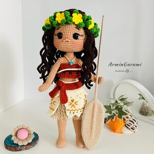 May include: Crocheted Moana doll with brown hair, a flower crown, a red and white top, a brown skirt, and a brown paddle. The doll is standing on a white surface. The text "ArminGurami Handmade" is visible in the background.