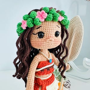 May include: Crocheted doll with brown hair, a flower crown, and a red and white dress. The doll is holding a brown crocheted paddle.