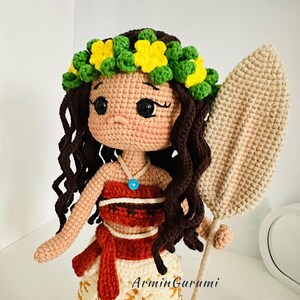 May include: Crocheted doll with brown curly hair, wearing a yellow flower crown, a red and white top, and a brown skirt. The doll is holding a brown crocheted paddle.