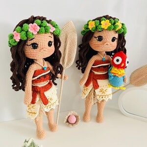 May include: Two crocheted dolls dressed as Moana from the Disney movie Moana. The dolls have brown hair, brown eyes, and are wearing a flower crown. One doll is holding a brown crocheted paddle and the other is holding a crocheted parrot.