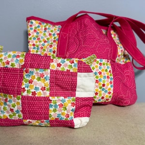 May include: A quilted tote bag and a matching rectangular zippered pouch featuring a patchwork design of bright pink fabric with white polka dots and a white fabric printed with small multicolored flowers, finished with two pink fabric handles.