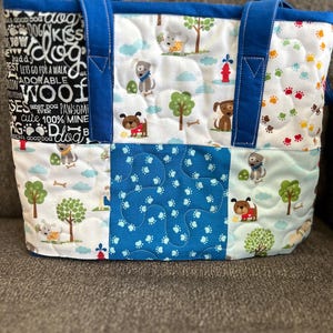 May include: A blue and white quilted tote bag with a dog theme. The bag has a blue handle and is decorated with paw prints, dogs, and trees. The text on the bag reads "A good dog" and "Best dog ever".
