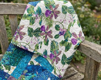 May include: A quilted fabric piece featuring a top section with a white background, purple orchid-like flowers, and green tropical leaves. The bottom section displays a patchwork design in shades of deep blue, teal, and purple with textured patterns.