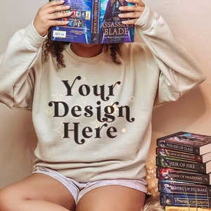 Puede incluir: Una sudadera beige con el texto "Your Design Here" en letras negras con detalles dorados. La sudadera la lleva puesta una persona sentada en una silla con una pila de libros delante.