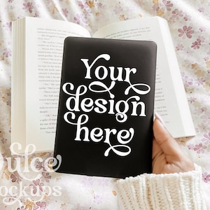 May include: A black notebook with the text "Your design here" in white lettering. The notebook is being held by a hand wearing a white sweater.