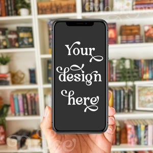 May include: A black smartphone with a dark screen displaying the text "Your design here" in white cursive font. The phone is held in a hand against a blurred background of a white bookshelf filled with books.