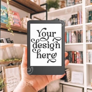 May include: A person is holding a black e-reader with a white screen that says "Your design here". The e-reader is in front of a bookshelf filled with books.