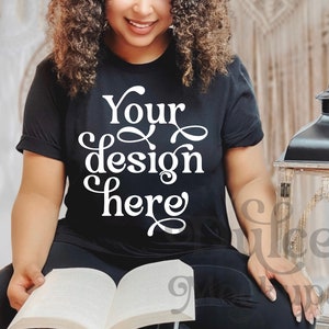 Pu&ograve; includere: Una maglietta nera con il testo "Your design here" in lettere bianche. La maglietta &egrave; indossata da una persona seduta e che legge un libro.