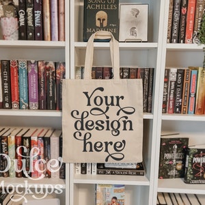 May include: A beige tote bag with black text that says "Your design here" on a bookshelf with books.