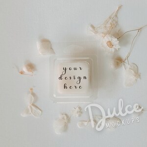 May include: A square, off-white product in a clear plastic container, with the text "your design here" in black script. The image is styled with dried flowers and the text "Dulce Mockups".
