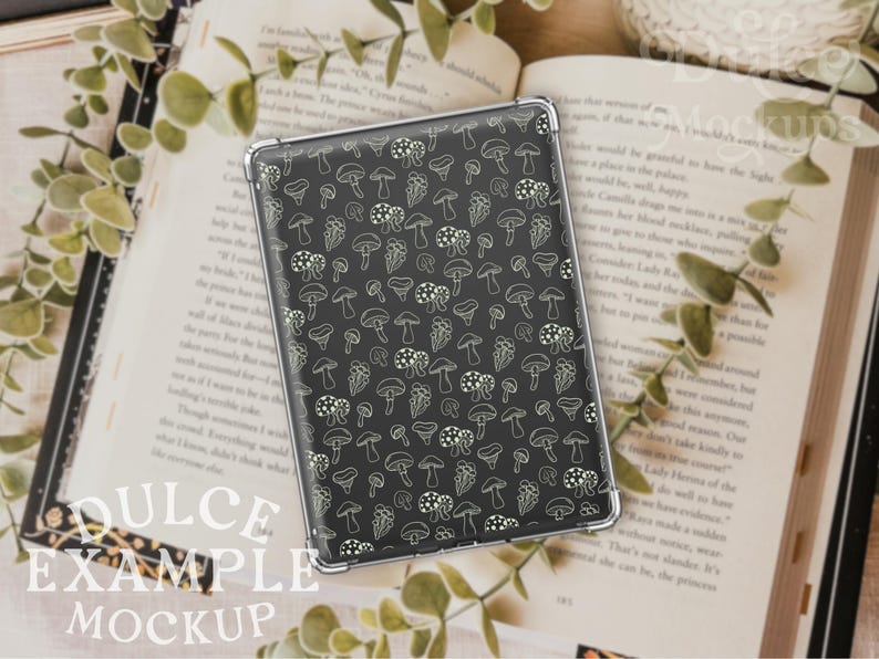 May include: A black phone case with a white mushroom pattern. The case is lying on a book with green leaves.