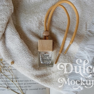 Puede incluir: Una pequeña botella de vidrio transparente con tapa de madera y cordón dorado, sobre una tela blanca. La botella tiene el texto "Your design here". El fondo incluye un libro y flores secas.