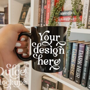 May include: A black coffee mug with a white handle and the text "Your design here" in a decorative font. The mug is being held by a hand in front of a bookshelf with books.