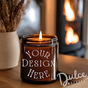 Op de afbeelding: Een bruine glazen kaars pot met een wit label waarop "YOUR DESIGN HERE" staat. De kaars is aangestoken en de vlam is zichtbaar. De kaars staat op een houten tafel voor een open haard.