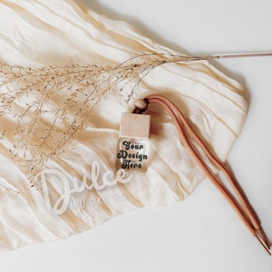 May include: A small glass bottle with a wooden cap and a brown cord. The bottle is filled with a clear liquid and has the text "Your Design Here" printed on it. The bottle is sitting on a white fabric background with a dried grass sprig.