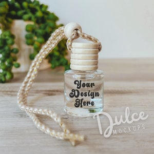 May include: Clear glass car air freshener bottle with a wooden cap and a beige rope. The bottle has the text "Your Design Here" printed on it. The background includes a white pot with a green plant and a wooden surface.